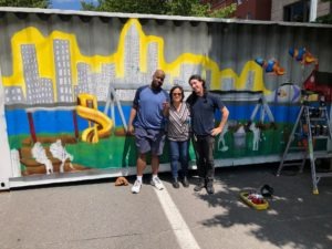 Three artists, Matthew Carroll, Fran Ledonia Flaherty, and Max Gonzales in front of a mural painted on the side of a shipping container
